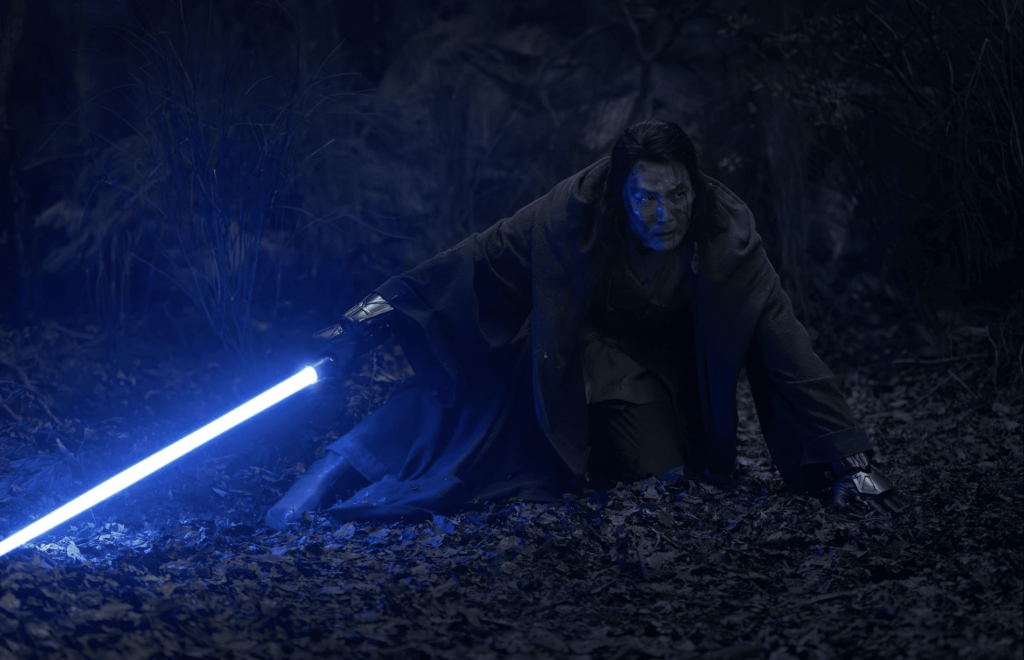 Man wielding blue lightsaber in dark, eerie forest.